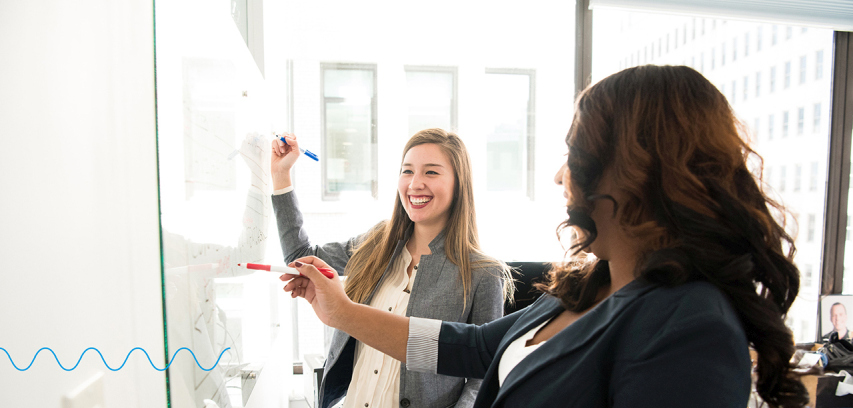 Business-agility-agile_orgs_photo_implementation_young_women_collaboration Two businesswomen laugh while collaborating on a dry erase board