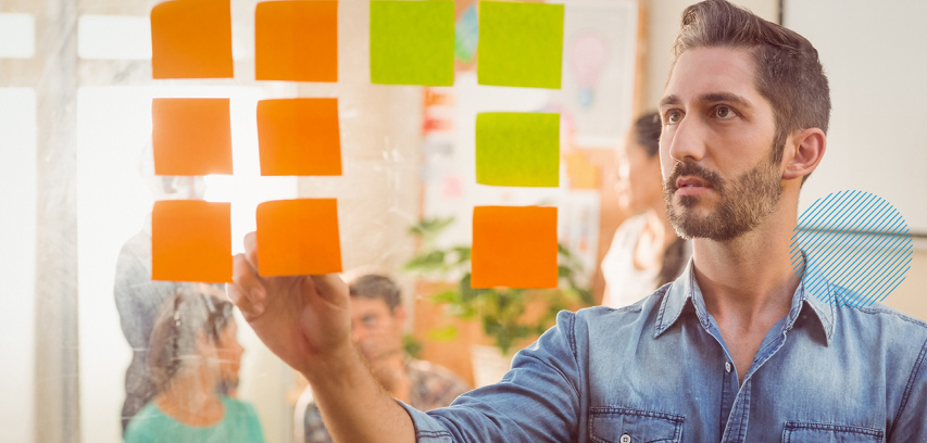 Business-agility-pillar-agile_orgs_photo_leadership_man_using_postit_notes A businessman places sticky notes on a glass wall and colleagues are seated in the background