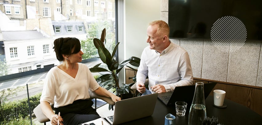 Business-agility-pillar_agile_orgs_photo_coaching_coach_talking_with_executive Two professionals sit in a conference room conversing in front of a large window