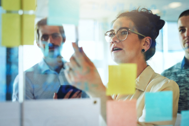 What-is-a-scrum-master-Who-are-scrum-masters Colleagues watch a scrum master place sticky notes on a glass work wall